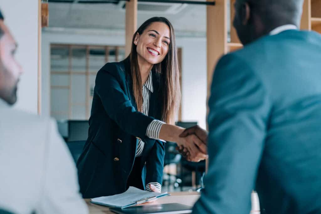 In an office setting, a woman in a suit smiles while shaking hands with a man, possibly discussing matters related to the Louisiana Public Defender's Office. Meanwhile, another individual is partially visible on the left.