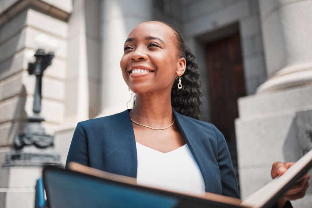 A woman in a suit smiles while holding a folder outside the Louisiana Public Defender's Office, a building with large stone columns.