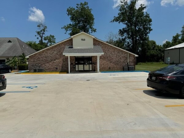 A brick building marked with the number 750, resembling the Louisiana Public Defender's Office, is nestled in a parking lot with a few cars. Trees stand tall against the backdrop of a clear sky.