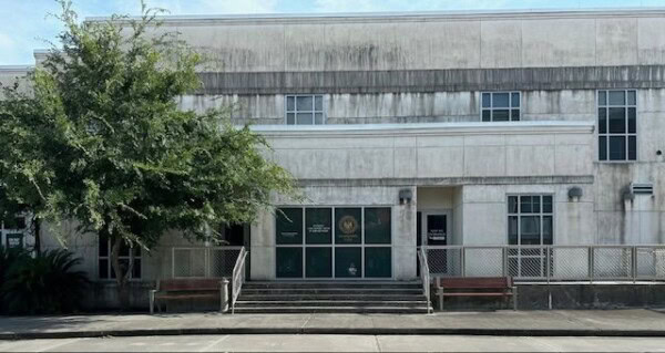A two-story, weathered concrete building houses the District 34, St. Bernard Parish location of the Louisiana Office of the State Public Defender. It features a central entrance with stairs and a ramp, flanked by two benches and with a tree on the left side.