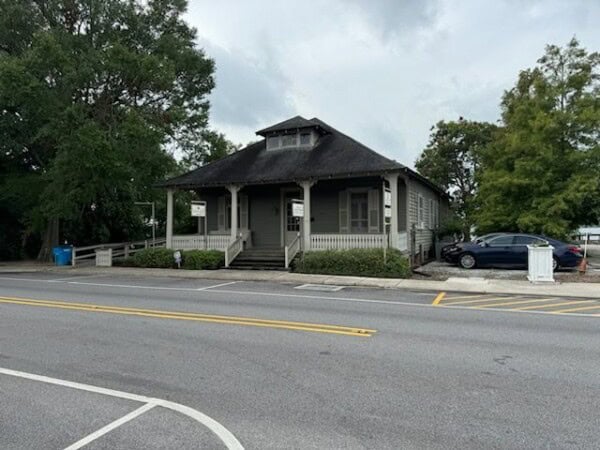 A small, grey, single-story building identified as the Louisiana Public Defender's Office features a covered porch and steps. It sits across a street with trees in the background, and cars are parked nearby.