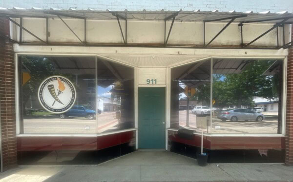Storefront with large windows and a green door in the center, reminiscent of the LAOSPD's open-door policy. A circular logo is displayed on the left window, while cars are reflected in the glass.