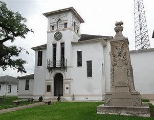 A white historic building with a clock tower and arched doorway, next to a tall stone monument on a grassy lawn, stands as home to the Louisiana Public Defender's Office.