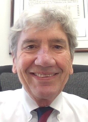 A smiling older man with gray hair, wearing a white shirt and tie, is sitting in an office chair with a document on the wall behind him. He exudes confidence as he represents the Louisiana Public Defender's Office.