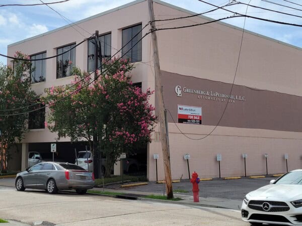 Two-story office building with "Greenberg & LaPeyronnie, LLC" sign, surrounded by parked cars and trees. A "For Sale" sign sits prominently in front, hinting at its potential conversion into the Louisiana Public Defender's Office or similar institution.