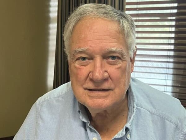 An older man with gray hair, wearing a light blue shirt, sits in a room associated with the Louisiana Public Defender's Office, as blinds cover the window behind him.