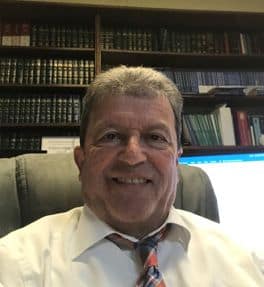 A person sitting in an office chair in front of bookshelves, wearing a white shirt and patterned tie, smiles at the camera, embodying the professionalism of the Louisiana Public Defender's Office.