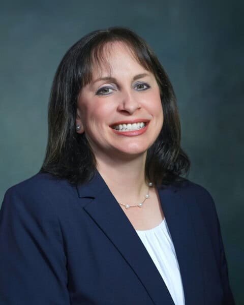 A woman with shoulder-length brown hair, wearing a navy blazer and white blouse, smiles confidently against a dark gray background, representing the Louisiana Public Defender's Office.