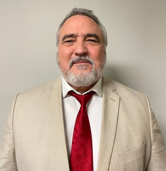 A man with gray hair and a beard, clad in a light-colored suit and red tie, stands poised against a plain, light backdrop. His presence embodies the dedication of the Louisiana Public Defender's Office.