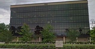 The six-story Louisiana Public Defender's Office features a dark glass exterior, surrounded by trees and shrubs under an overcast sky.
