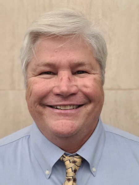 A person with grey hair is smiling, wearing a light blue shirt and patterned tie, set against a neutral background, reminiscent of a professional portrait one might find at the Louisiana Public Defender's Office.