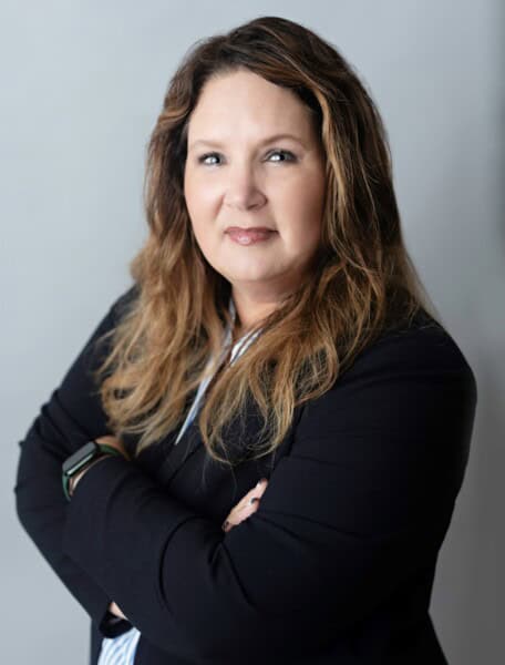 A person with long wavy hair, wearing a black jacket, stands with arms crossed against a plain background, exuding an air of confidence often seen in the dedicated members of the Louisiana Public Defender's Office.