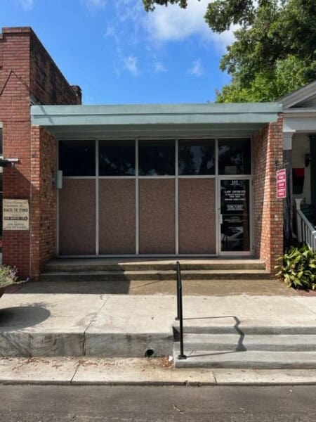 The Louisiana Public Defender's Office is situated in a brick building with large windows and a glass door, complemented by a small set of stairs and a handrail lining the sidewalk.