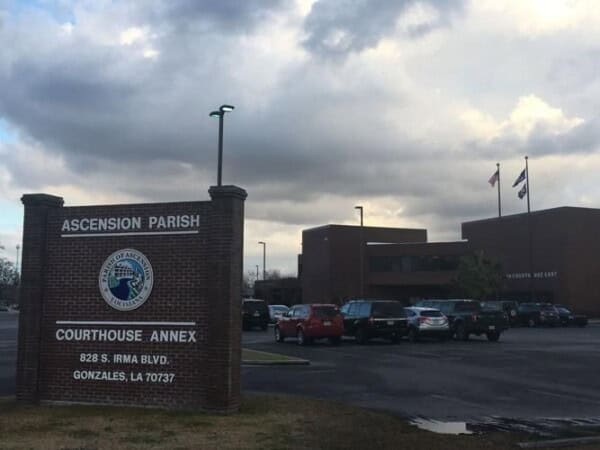 A brick sign for the Ascension Parish Courthouse Annex stands in a parking lot, with a building and flagpoles under a cloudy sky. Nearby, the Louisiana Public Defender's Office operates to ensure justice is served.