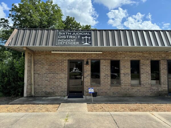 A brick building with a sign reading "Sixth Judicial District Indigent Defender" and scales of justice icon above the entrance marks the outdoor view, representing the Louisiana Public Defender's Office mission.